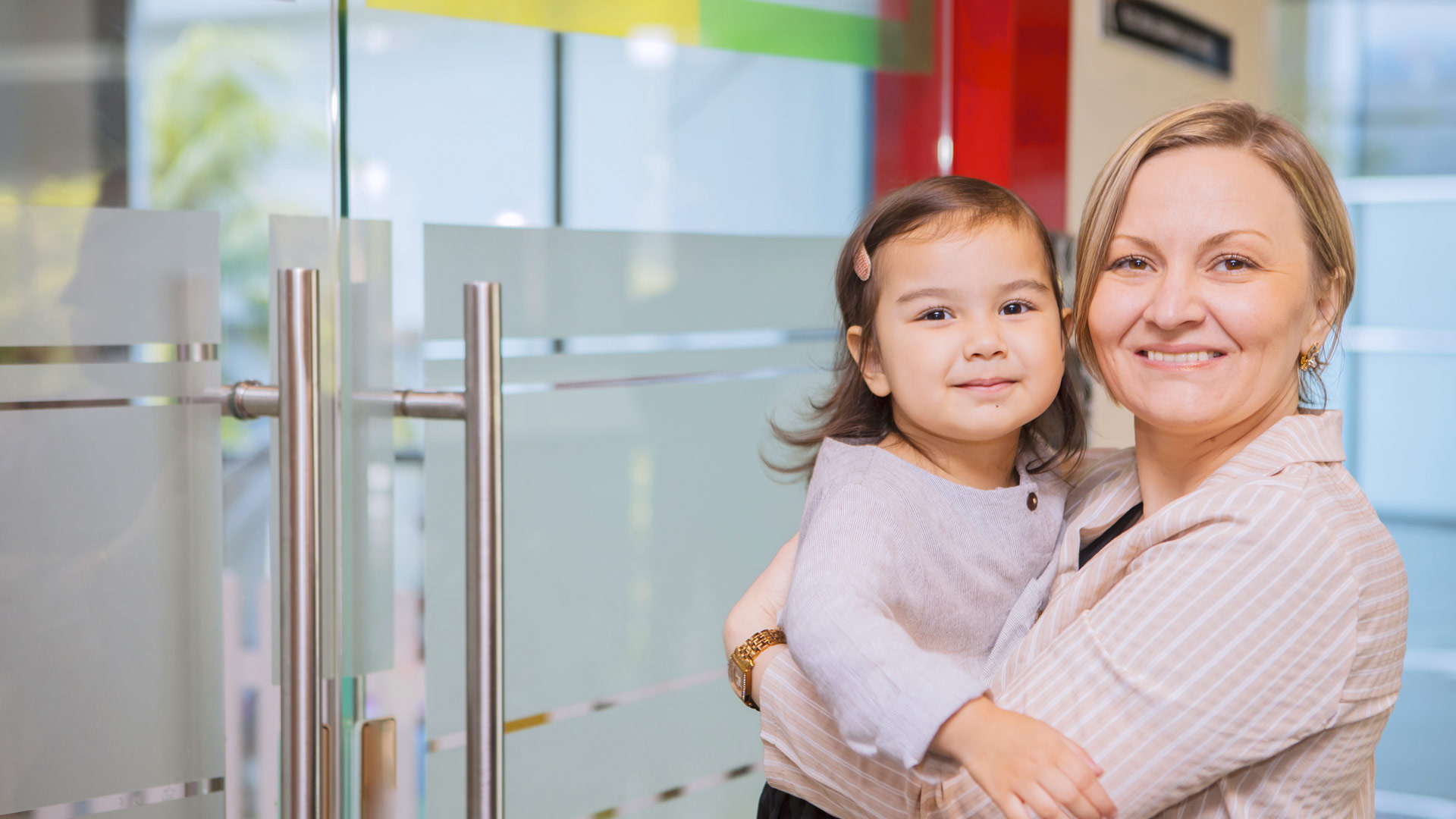 Une maman et sa fille sourient devant la porte vitrée d'un service de garde