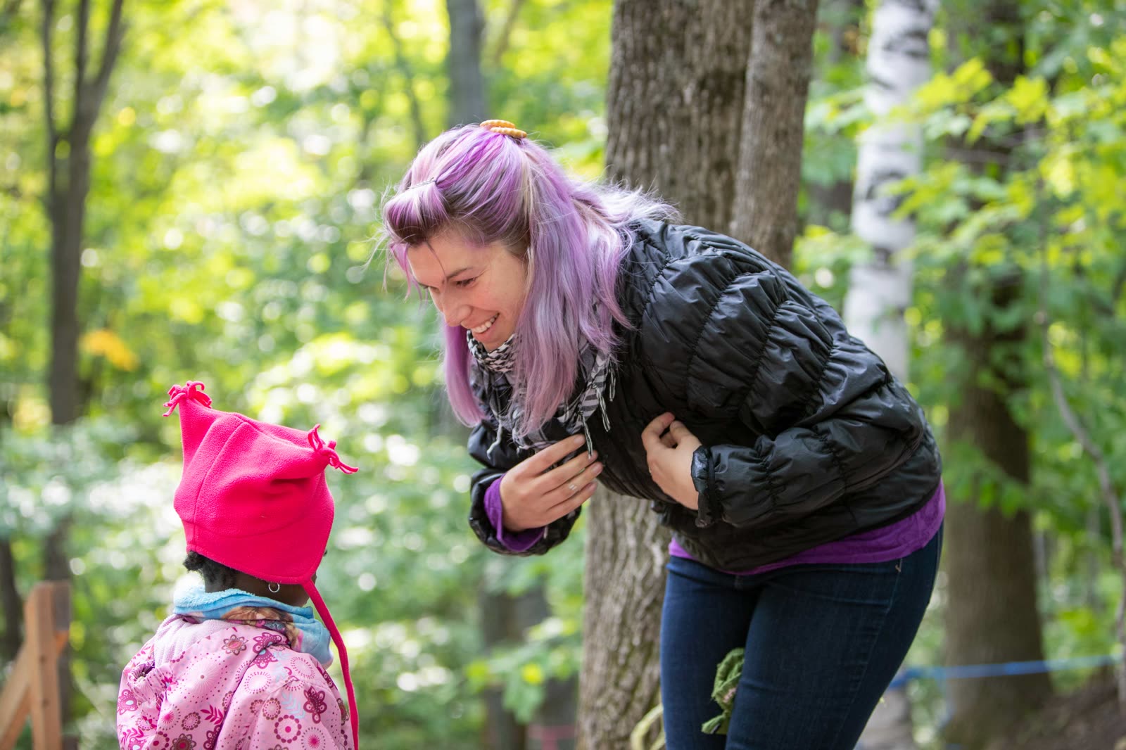Une éducatrice se penche pour écouter une enfant en tuque rose pendant une sortie en forêt